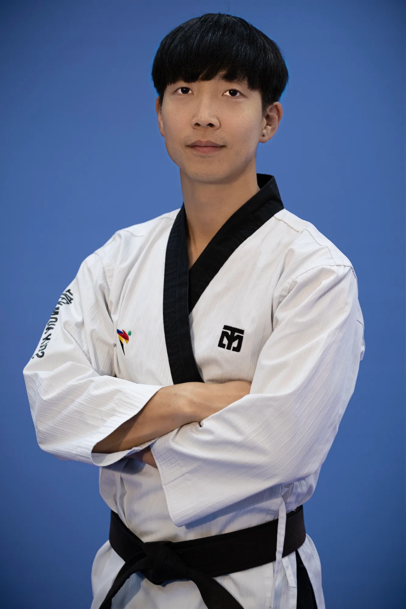 李周原师范身着白色道服、系黑带，双臂交叉站在蓝色摄影背景前